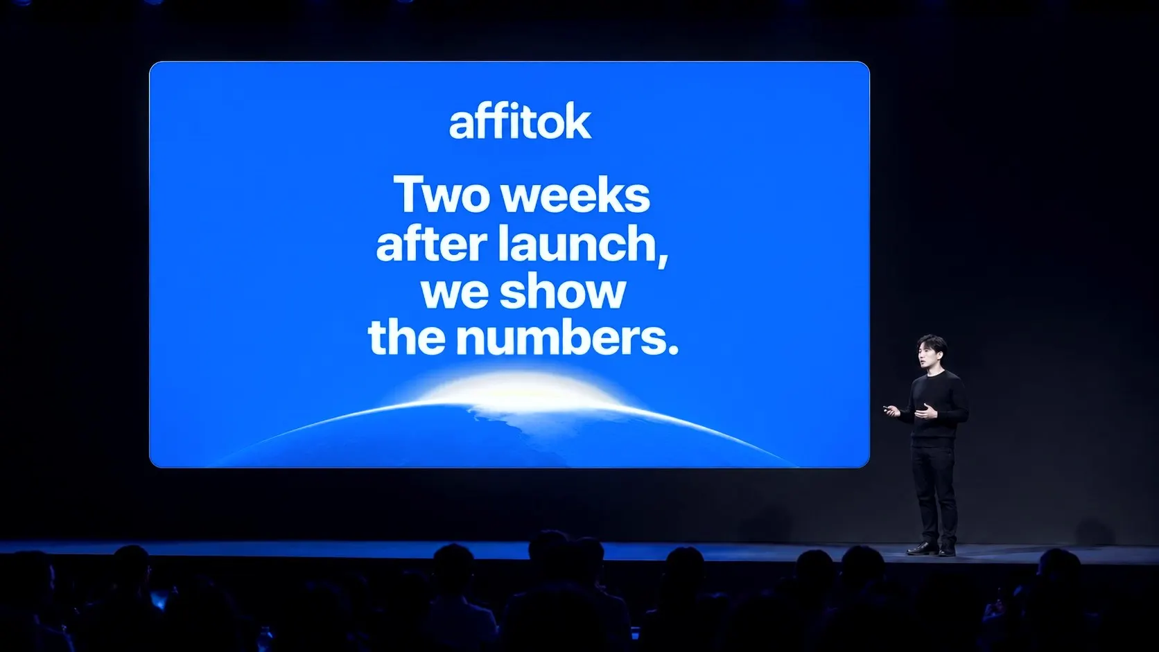 Conference stage: the big screen shows a blue slide with the affitok wordmark, Two weeks after launch, we show the numbers., over a rising sun and the blue earth; a presenter in black stands at the right
